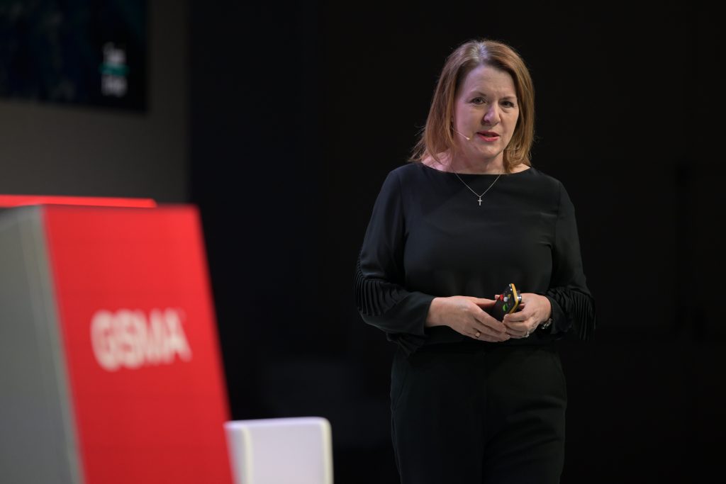 A woman with shoulder-length light brown hair, wearing a black long-sleeve top and pants, stands on stage holding a small device. She faces the audience, smiling slightly. In the foreground, there is a red podium with "GSMA" written on it.