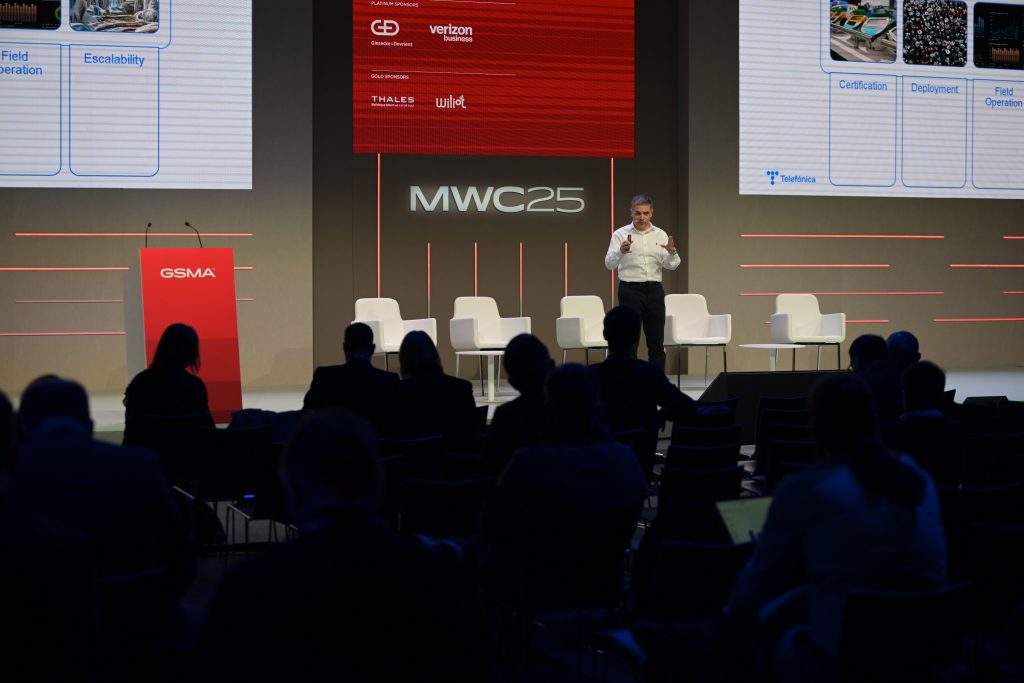 A speaker stands on stage at MWC25 before a seated audience. He holds a microphone, presenting near large red and white screens showing logos and slides. Empty chairs and a GSMA podium are visible, with the audience mostly in silhouette.