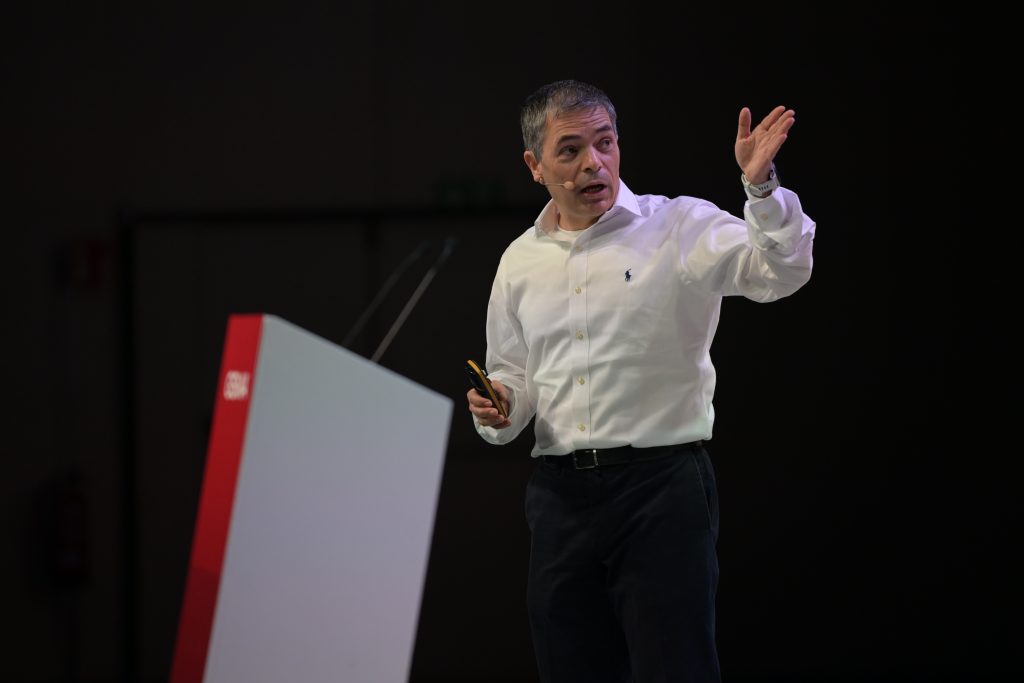 A man in a white shirt and dark pants stands on stage, gesturing with his right hand while holding a device in his left. He faces an audience, speaking near a podium with a red stripe, against a dark background.