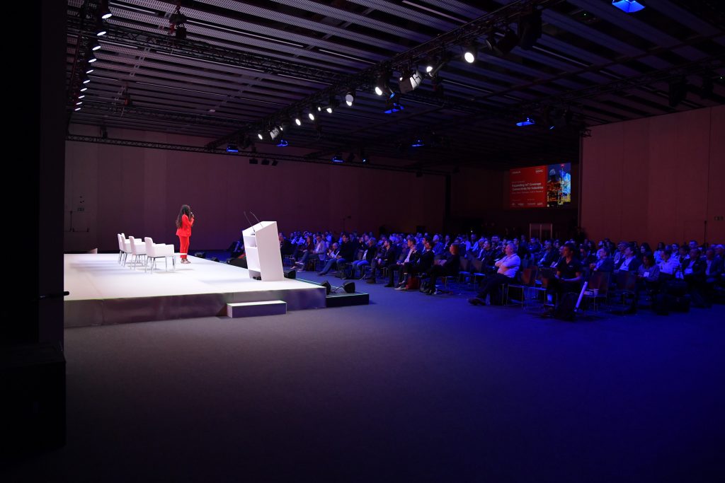 A woman in a red outfit stands on a brightly lit stage, speaking to a large seated audience in a dimly lit conference hall. Four empty white chairs and a podium are on stage; a red-lit screen is visible in the background.