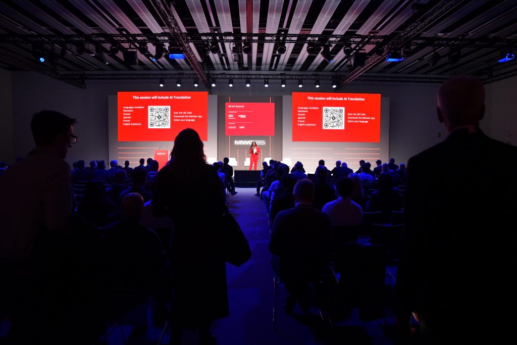 A speaker stands on a lit stage in front of an audience in a dark room. Red screens behind display text, a QR code, and event information. People are seated and a few are standing, silhouetted against the light from the stage and screens.