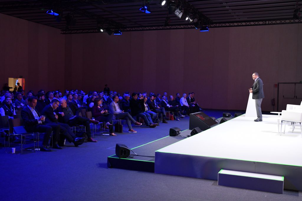 A man in a suit stands on a brightly lit stage addressing an audience seated in rows at a large conference. The audience watches attentively. The background is dim, highlighting the speaker and white chairs on stage.