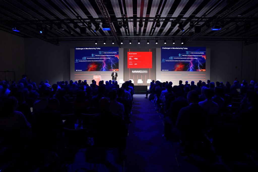 A large audience sits in a dark conference room facing a brightly lit stage where a speaker presents in front of two large screens displaying slides on manufacturing challenges at MWC23. The stage backdrop is mainly blue and red, with white text and graphics.