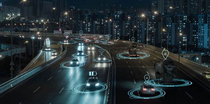 A nighttime city highway with several cars and trucks driving. Digital blue icons, such as Wi-Fi and location pins, are superimposed over vehicles, indicating connectivity and communication between them, with tall buildings and city lights in the background.