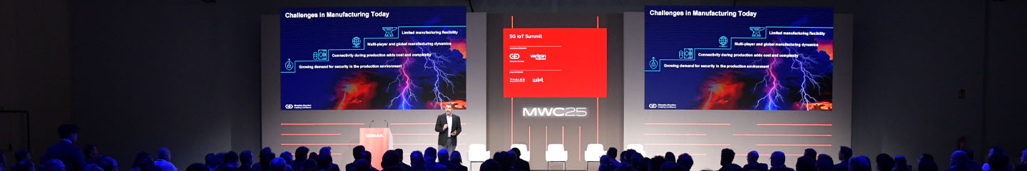 A speaker stands on stage at MWC25 in front of two large screens showing a blue and red presentation with lightning graphics and text about manufacturing challenges, facing an audience in a dark auditorium.