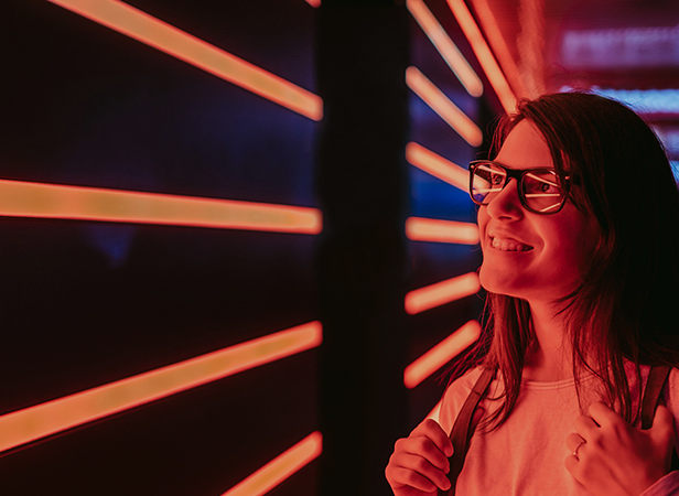 A young woman with long hair and glasses smiles while looking ahead. Sporting a white shirt and backpack, she stands beside neon orange-red lights—a vibrant, futuristic scene that reflects the innovative spirit of the 5G IoT Strategy Group.