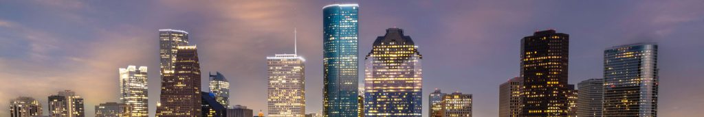 A panoramic view of a city skyline at dusk, featuring tall modern skyscrapers with illuminated windows and building outlines against a purple and pink cloudy sky. The tallest building is centrally located and brightly lit.