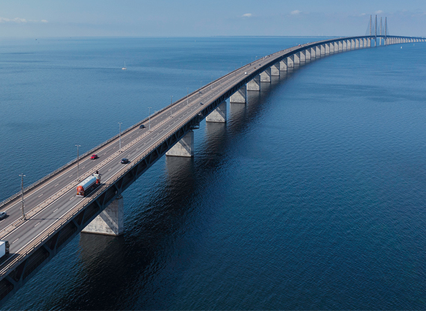 A long bridge with multiple lanes for vehicles extends over calm blue water, supported by numerous concrete pillars. As cars and trucks travel, the seamless connectivity of Mobile IoT (LPWA) roaming ensures smooth operations under the clear sky.