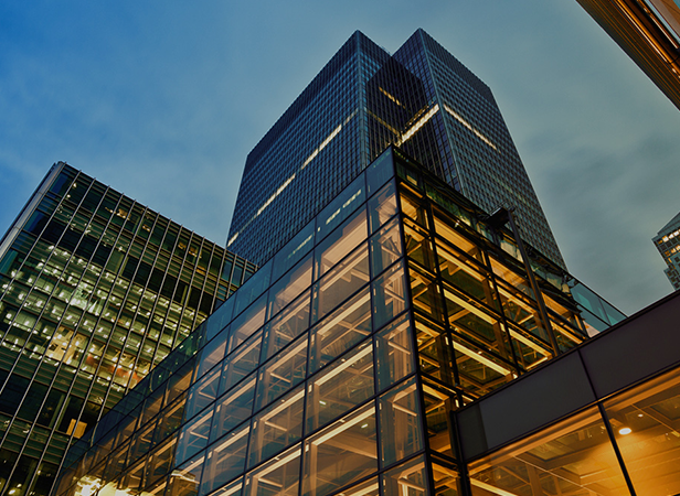 A low-angle view of modern glass office buildings at dusk, where the GSMA IoT Marketing Group's headquarters glow with warm lights. Reflective windows and illuminated interiors highlight the height and sleek lines against the cool blue evening sky.
