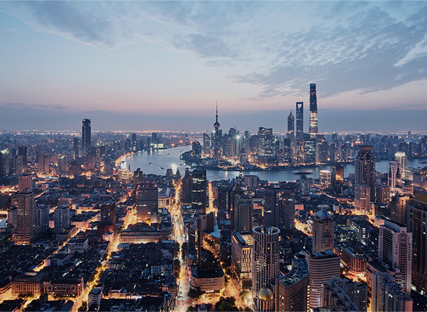 A panoramic view of a modern city at dusk, where tall skyscrapers and illuminated streets showcase a dense urban landscape infused with massive IoT, as the city lights begin to glow along the winding river beneath a blue and orange sky.