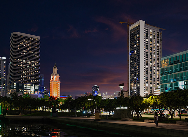 A cityscape at dusk shows tall modern buildings with 5G IoT-enabled illuminated windows, a historic beige tower with a lit top, palm trees lining the street, and a purple-orange sky. The edge of a waterfront is visible in the foreground with reflections on the water.