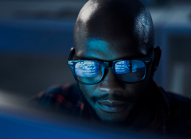 A man wearing glasses looks at a computer screen in a dimly lit room. Blue light reflects text from the screen onto his glasses, suggesting he is focused on IoT security or coding digital data with intense concentration.