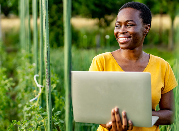 A smiling person in a yellow shirt stands outdoors holding a laptop, surrounded by green plants and vertical supports in an IoT environment, suggesting smart gardening on a warm, sunny day. The lush background is blurred for effect.