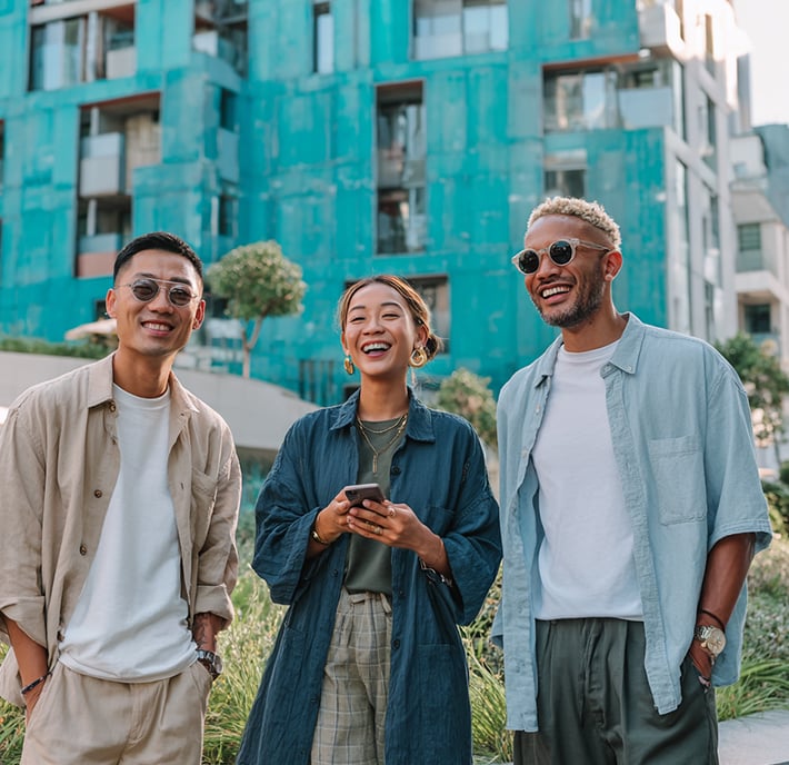 Three people smiling outdoors in front of a modern teal building, one holding a phone, dressed in casual clothes—perhaps discussing the latest LPWAN technology.