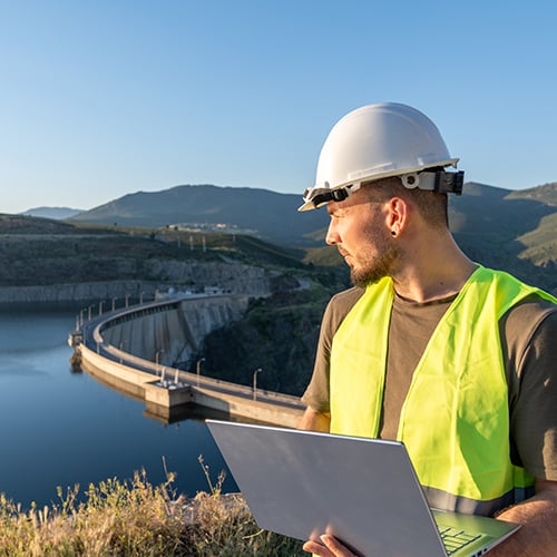 LPWAN Engineer in a hard hat and safety vest holds a laptop while inspecting a dam by a reservoir.