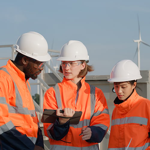 Three workers in orange safety gear and helmets discuss lpwan plans outdoors near wind turbines.