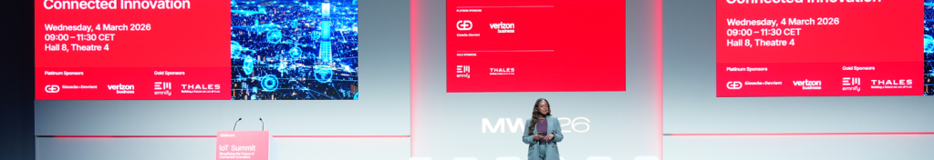 A woman speaks on stage at a tech conference with large red presentation screens behind her.