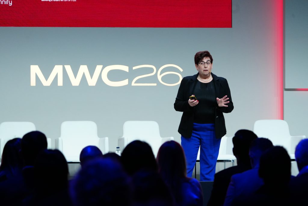 A person speaks on stage at MWC 26, with an audience watching and empty chairs behind them.