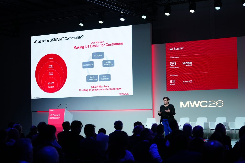 A speaker presents a slide about the GSMA IoT Community at MWC 26 to an audience in a conference room.