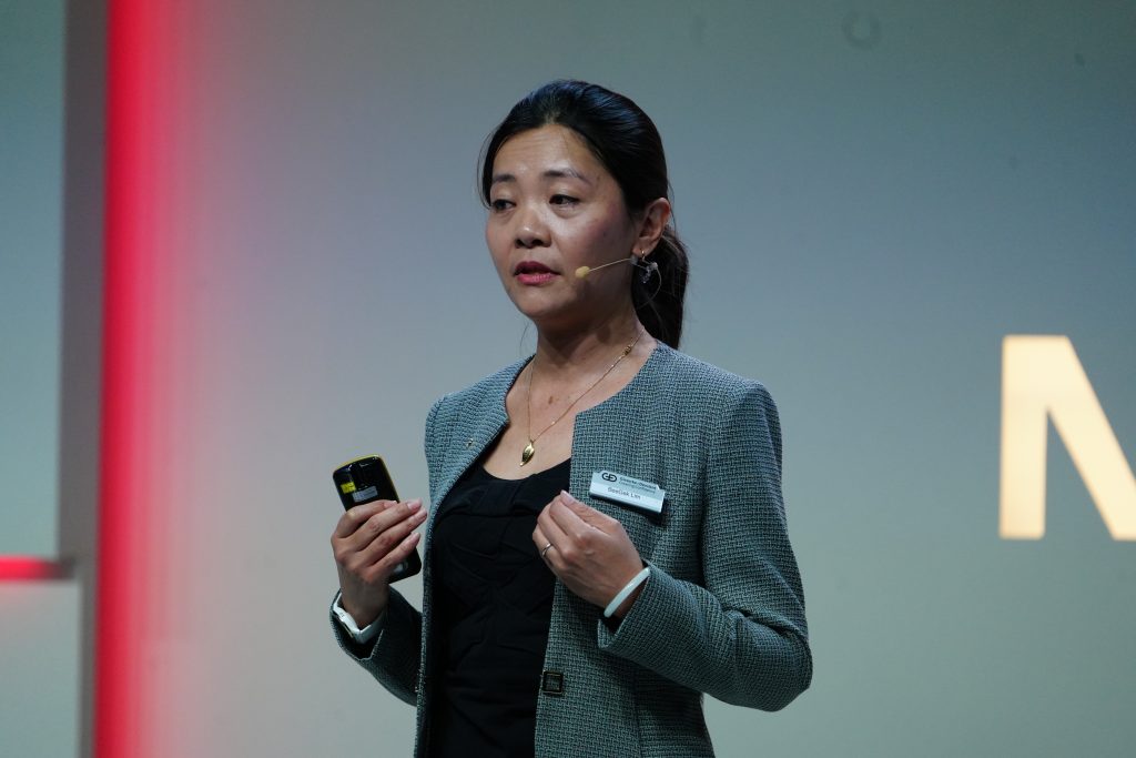 Woman in a gray blazer speaks on stage with a microphone headset, holding a device in her hand.