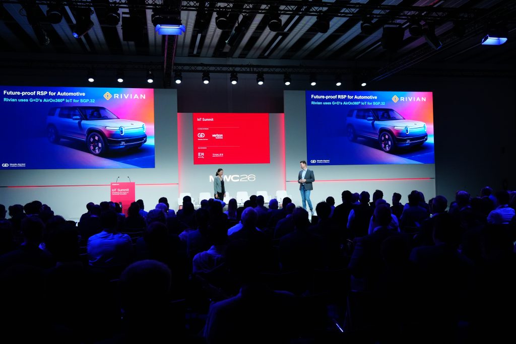 Two presenters on stage talk about RIVIAN automotive tech with a truck image on a large screen at a tech conference.