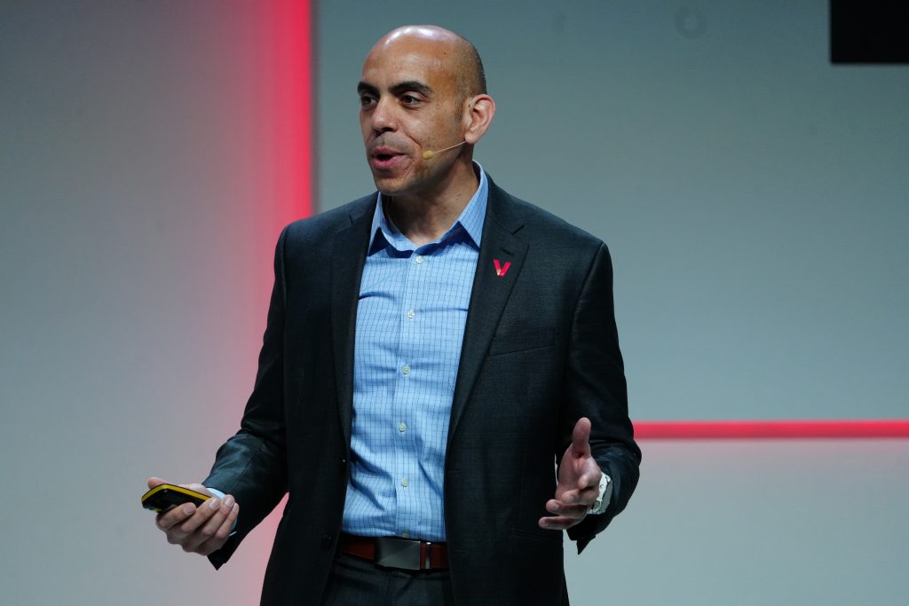 A man in a suit speaks on stage, holding a phone, with a red light accent in the background.