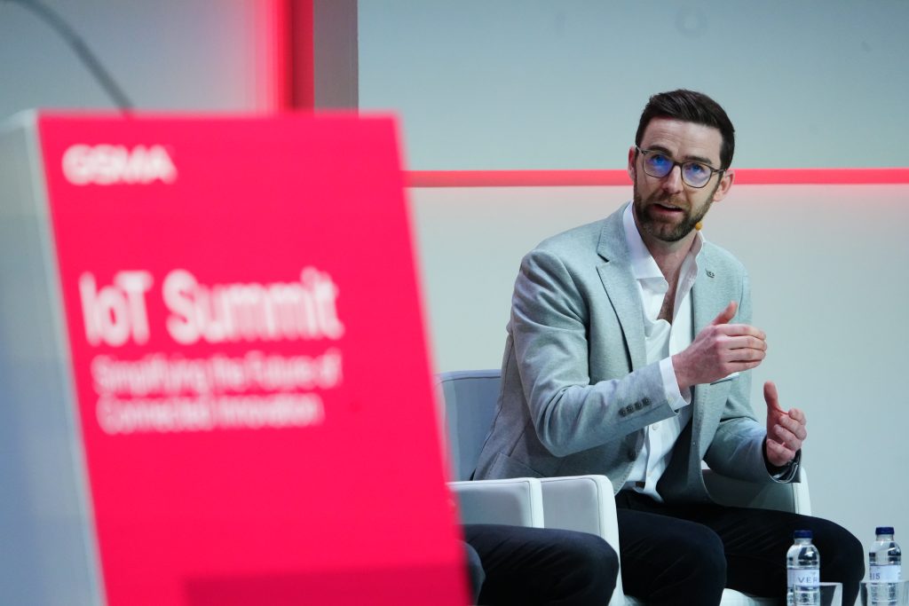 A man in glasses speaks at the IoT Summit, gesturing with his hands while seated on stage.