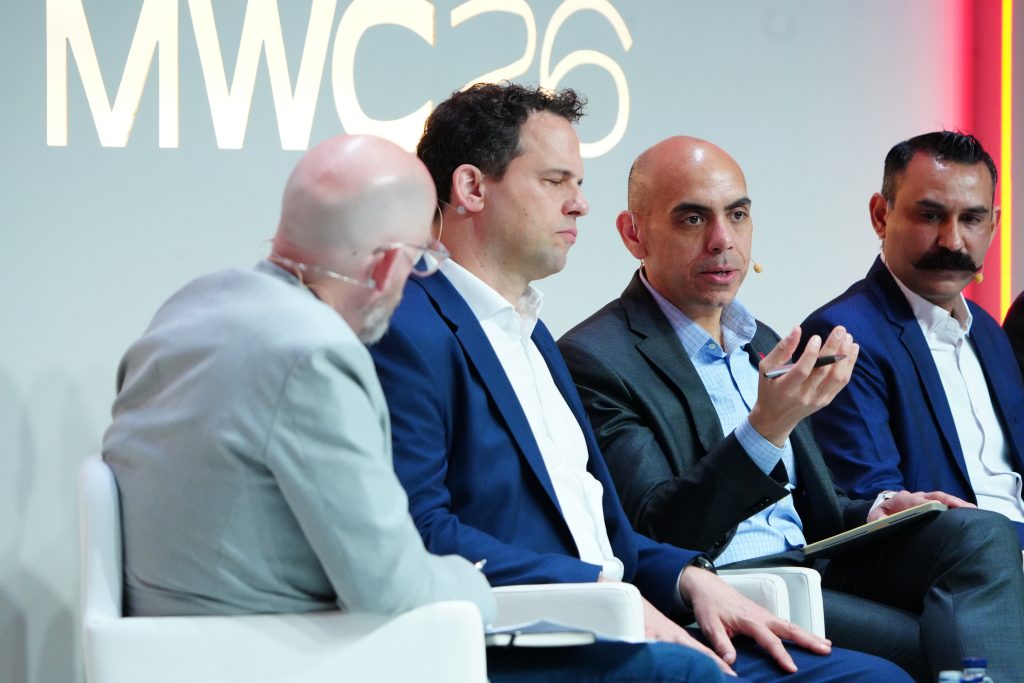 Four men in suits sit and talk on a panel discussion at an event with “MWC23” displayed in the background.