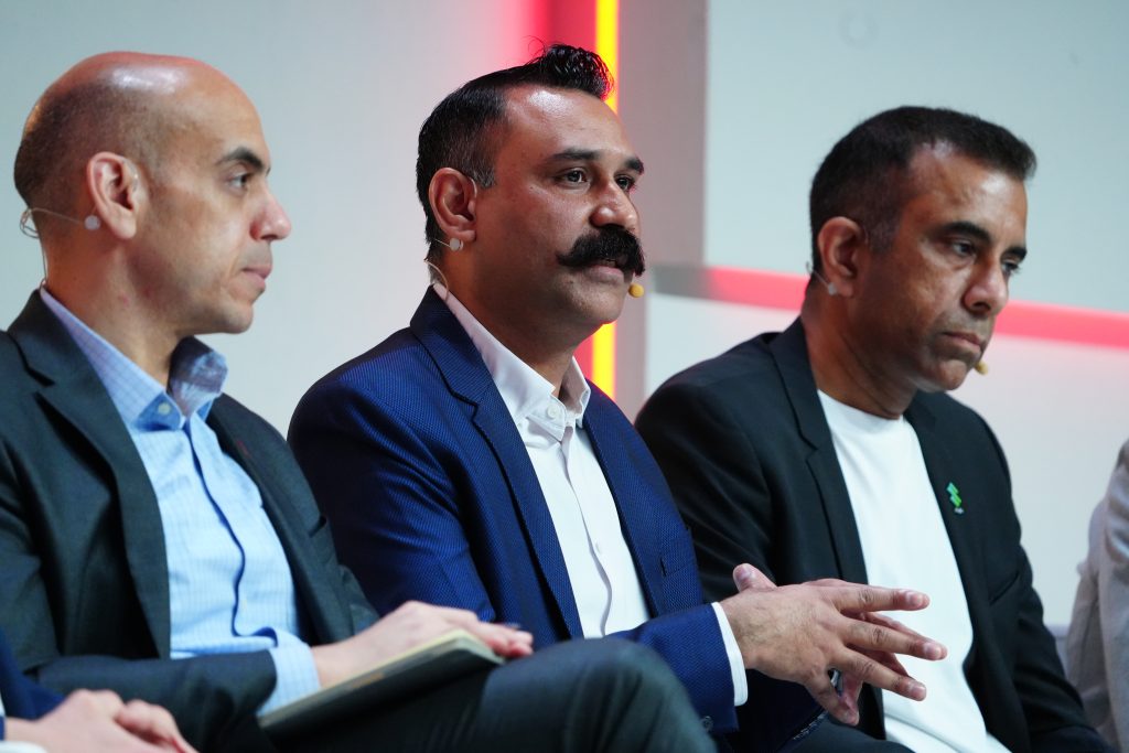 Three men in suits sit side by side, two listening while the man in the center speaks and gestures with his hands.