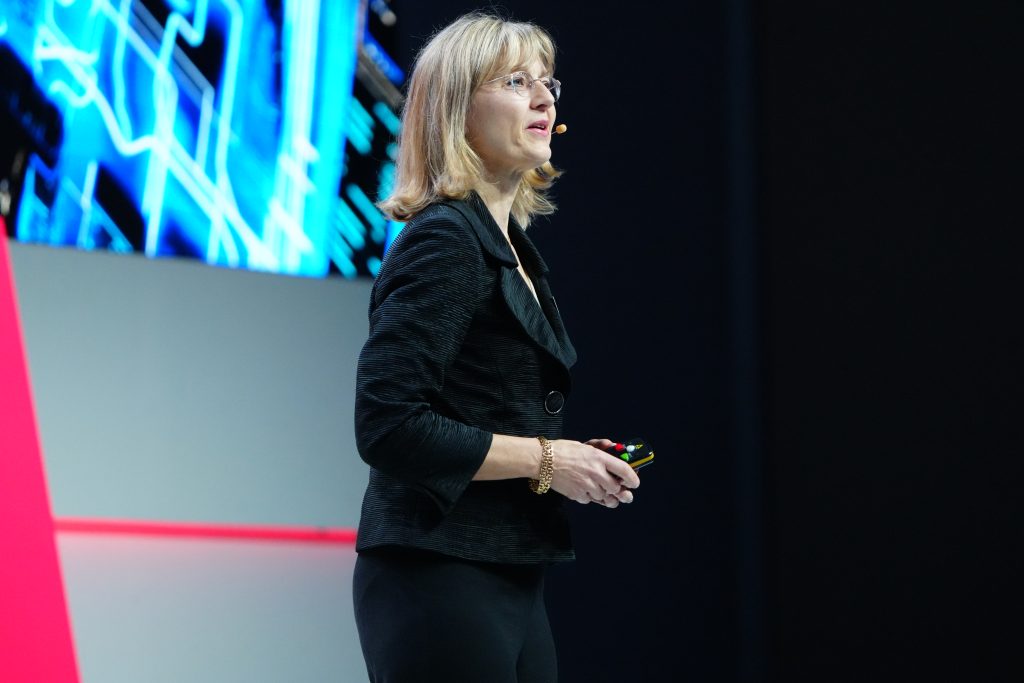 A woman speaks on stage with a headset microphone, standing near a screen displaying blue digital graphics.
