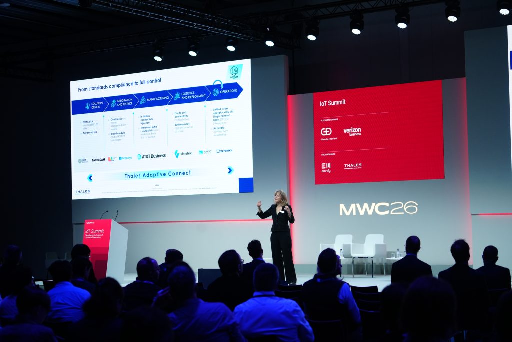 A woman presents on stage at a tech conference; attendees watch a slide and red display reading "MWC26 IoT Summit.
