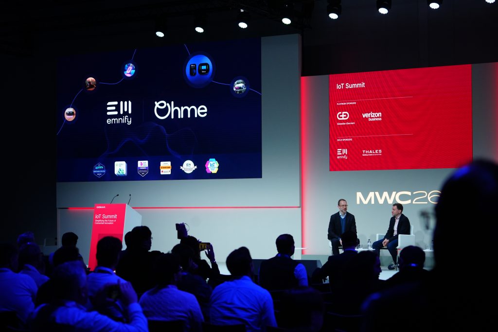 Two men speak onstage at a tech conference with a large screen displaying company logos behind them.