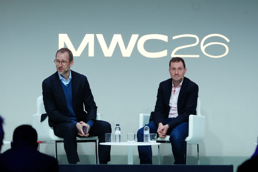 Two men seated on stage at MWC26 event, with microphones and water bottles on a table between them.