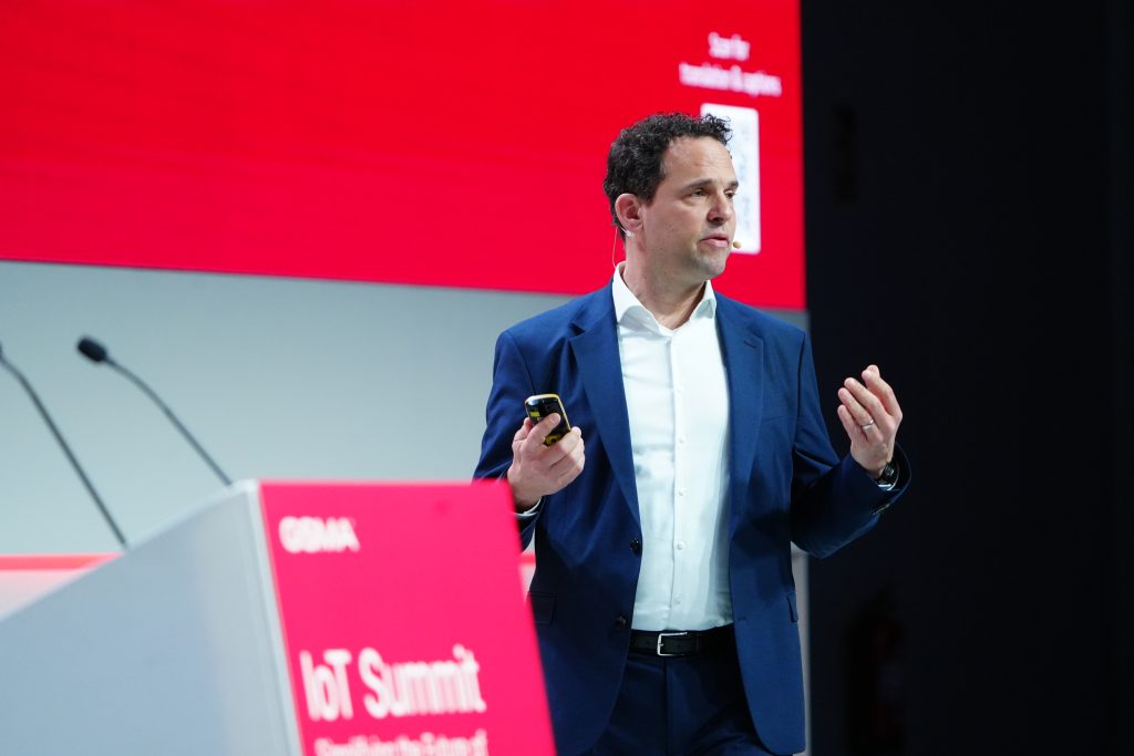 A man in a suit speaks on stage at the IoT Summit, holding a device, with a red screen behind him.