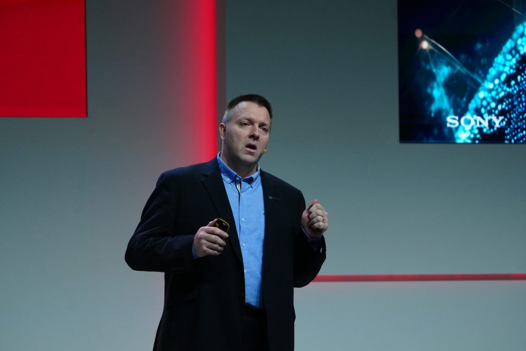 A man in a suit gives a presentation on stage with a screen displaying "Sony" in the background.