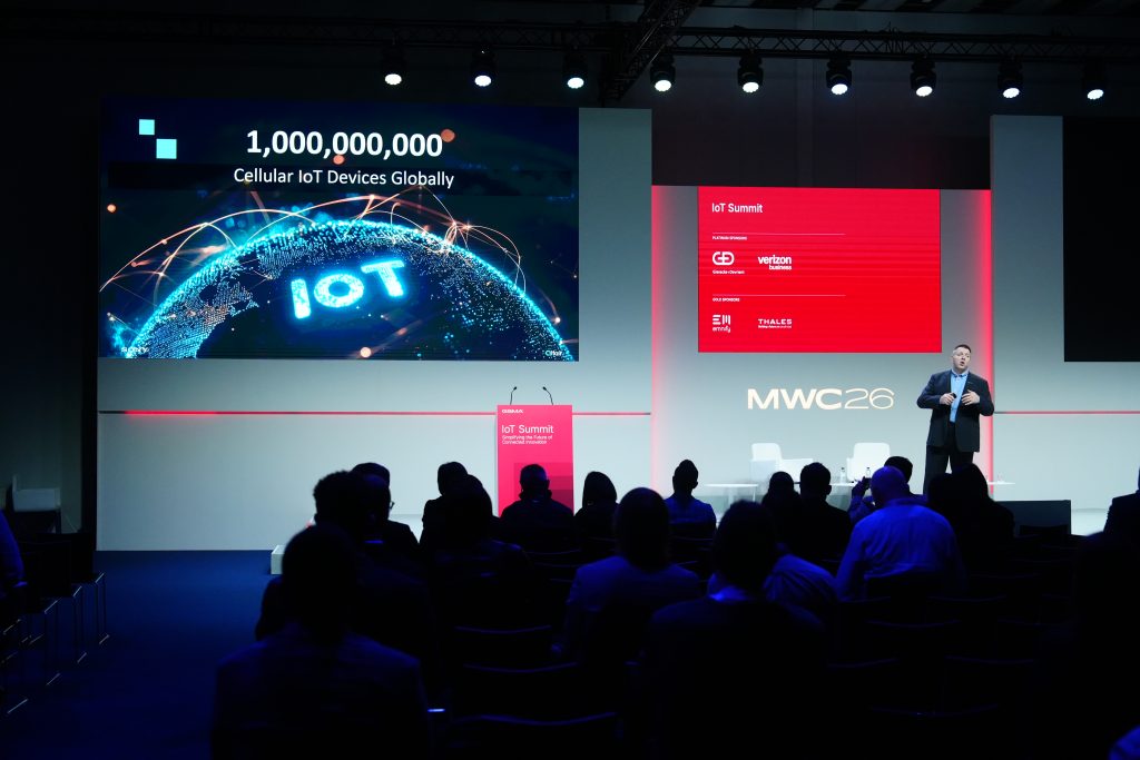 A speaker presents on stage about global IoT device growth at MWC26, with attendees seated in the audience.