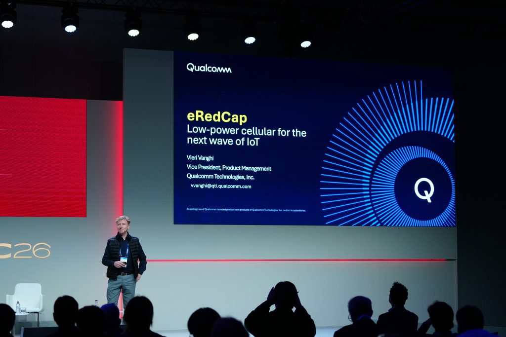 A speaker stands on stage beside a screen presenting "eRedCap: Low-power cellular for the next wave of IoT" by Qualcomm.