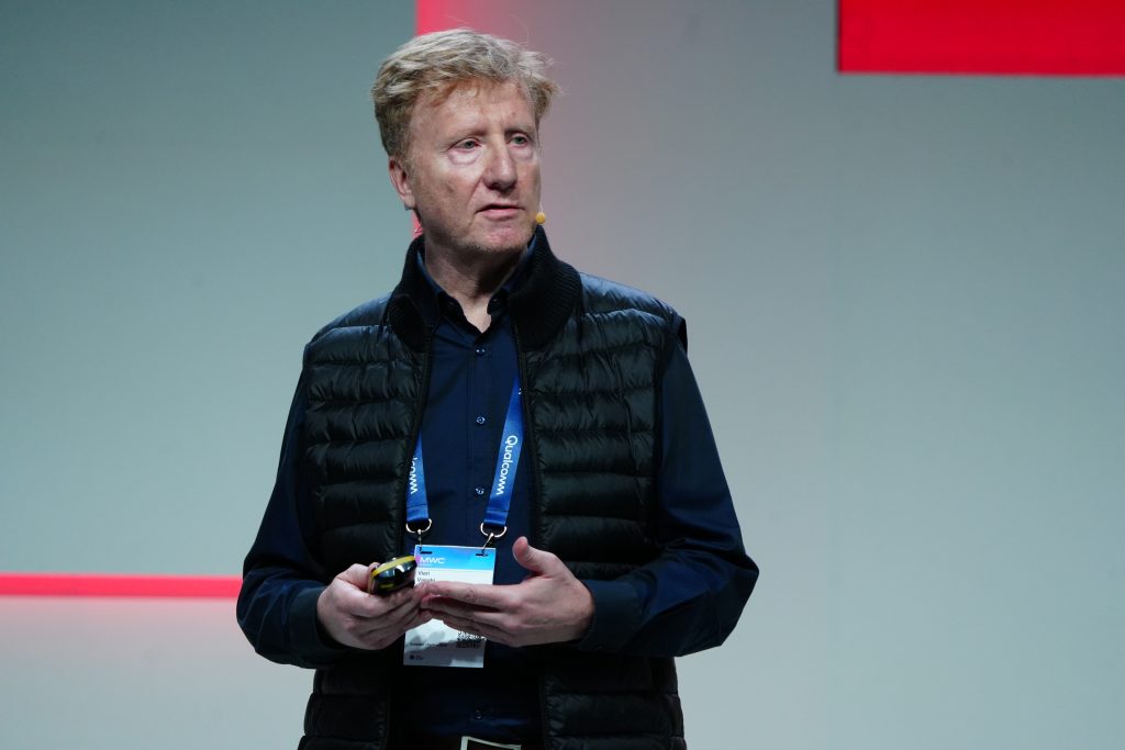 A man with a headset speaks onstage, wearing a black vest and holding a device, with a badge around his neck.