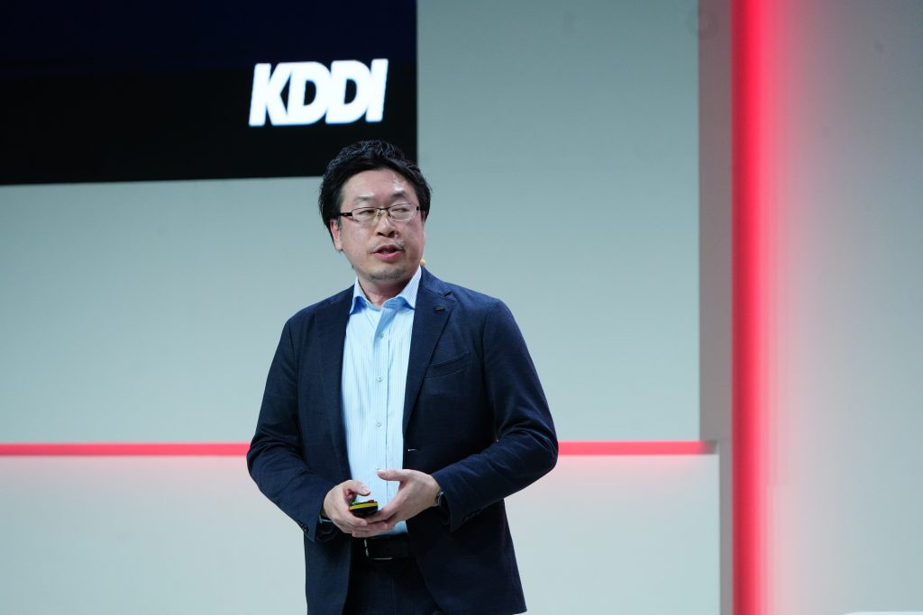 A man in a suit speaks on stage near a wall with the KDDI logo and red accent lighting.
