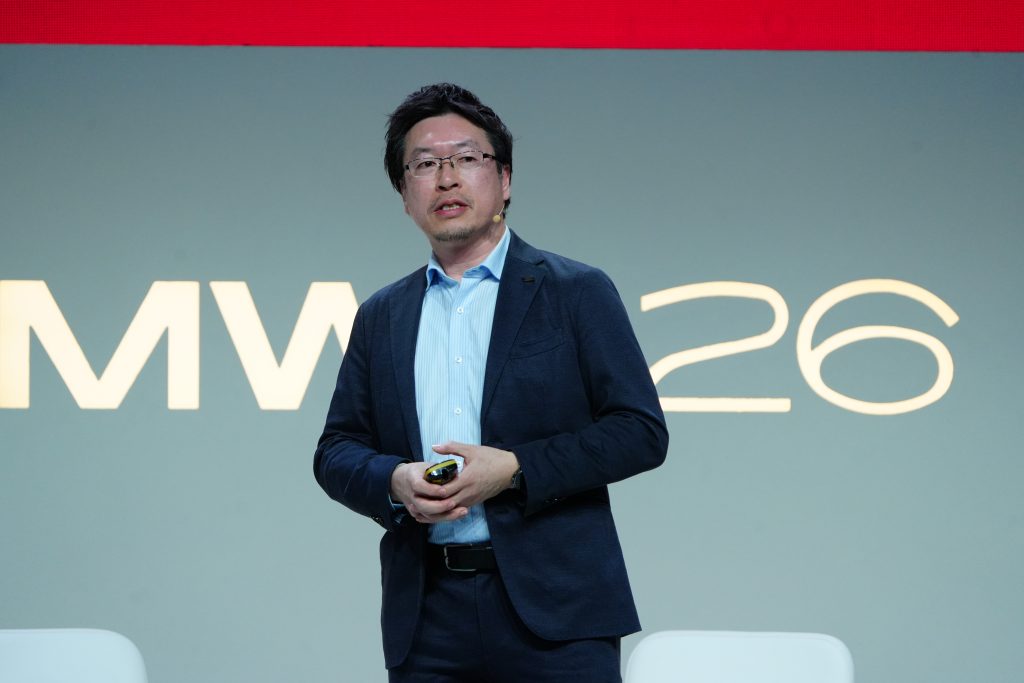 Man in a suit speaking on stage with "MWC 26" displayed in large letters behind him.