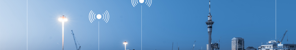 City skyline at dusk with cranes, tall buildings, and streetlights; white wireless signal icons are digitally overlaid above the lights, suggesting technology or connectivity in the urban landscape. The sky is clear and blue, transitioning from daylight to evening.