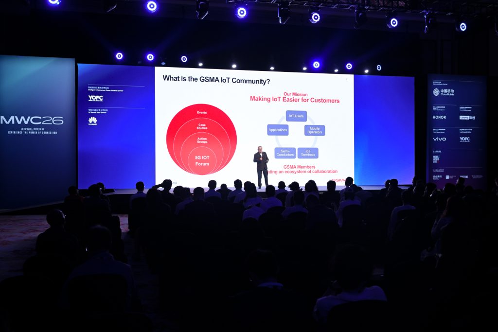 A conference room with an audience facing a large screen displaying a GSMA IoT Community presentation. The slide shows red diagrams and text about collaboration, surrounded by blue-lit stage walls featuring sponsor logos and the event name "MWC26.