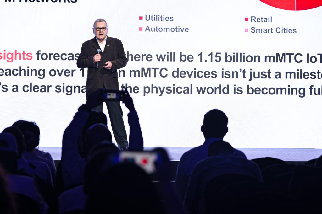 A man in a suit stands on stage holding a mic, presenting in front of a screen with bold text and graphs. The audience sits in front, some taking photos. The slide mentions IoT, mMTC devices, utilities, automotive, retail, and smart cities.