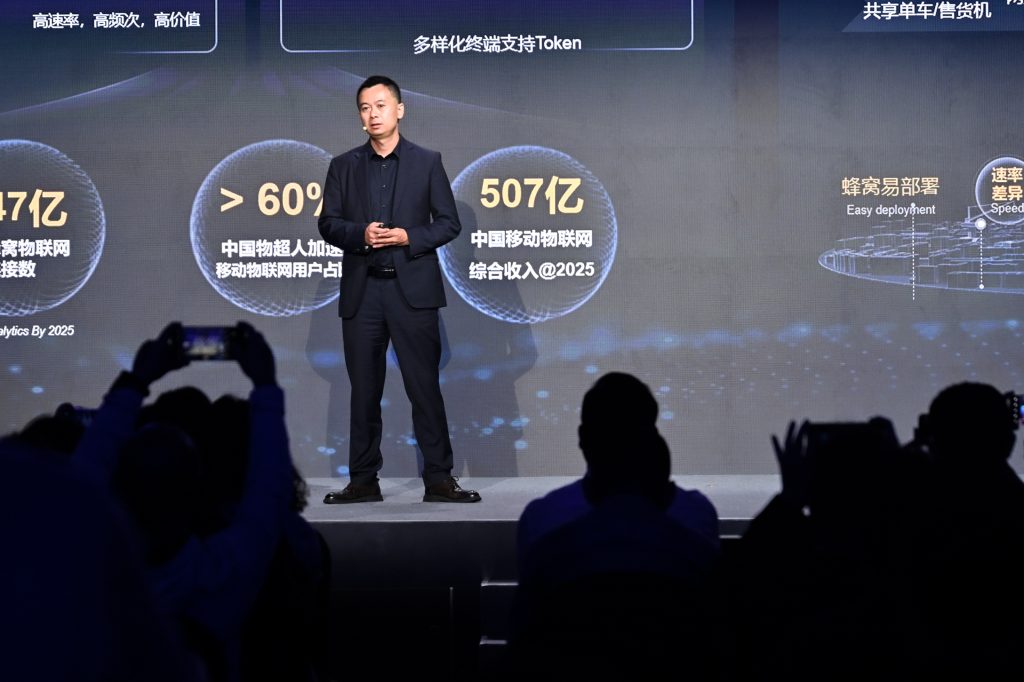 A man in a dark suit stands onstage against a black backdrop with Chinese text, numbers, and diagrams. Audience members in the foreground watch and take photos. The scene appears to be a business or tech presentation or conference.
