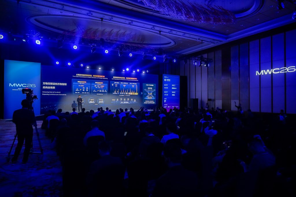 A large audience sits in a dark conference hall facing a brightly lit stage with multiple presentation screens displaying charts and text. The event features blue lighting, a high ceiling, and "MWC23" branding is visible on a wall and a screen.