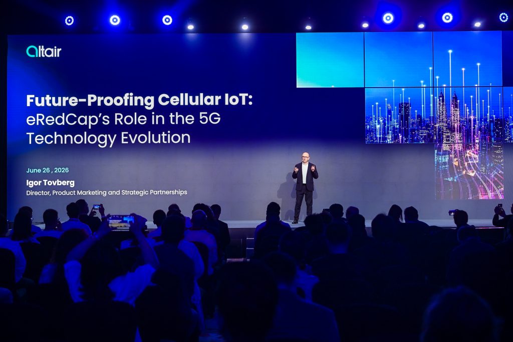 A man stands onstage presenting to an audience. Behind him, a large screen displays the title “Future-Proofing Cellular IoT: eRedCap’s Role in the 5G Technology Evolution,” along with his name, “Igor Tovberg,” and a graphic of a digital cityscape.