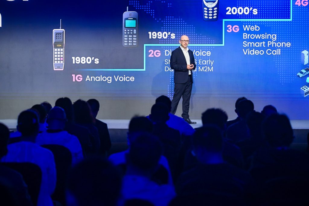 A man in a suit speaks onstage before an audience. Behind him is a large screen displaying the evolution of mobile phones and networks from 1G in the 1980s to 4G, with images of phones and key features for each generation.