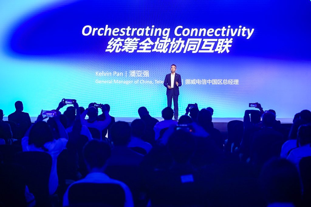 A man in a suit stands on stage under the text “Orchestrating Connectivity” in English and Chinese, addressing a seated audience. Some attendees hold up phones to record. Blue and white lights illuminate the stage, creating a modern, tech-focused atmosphere.