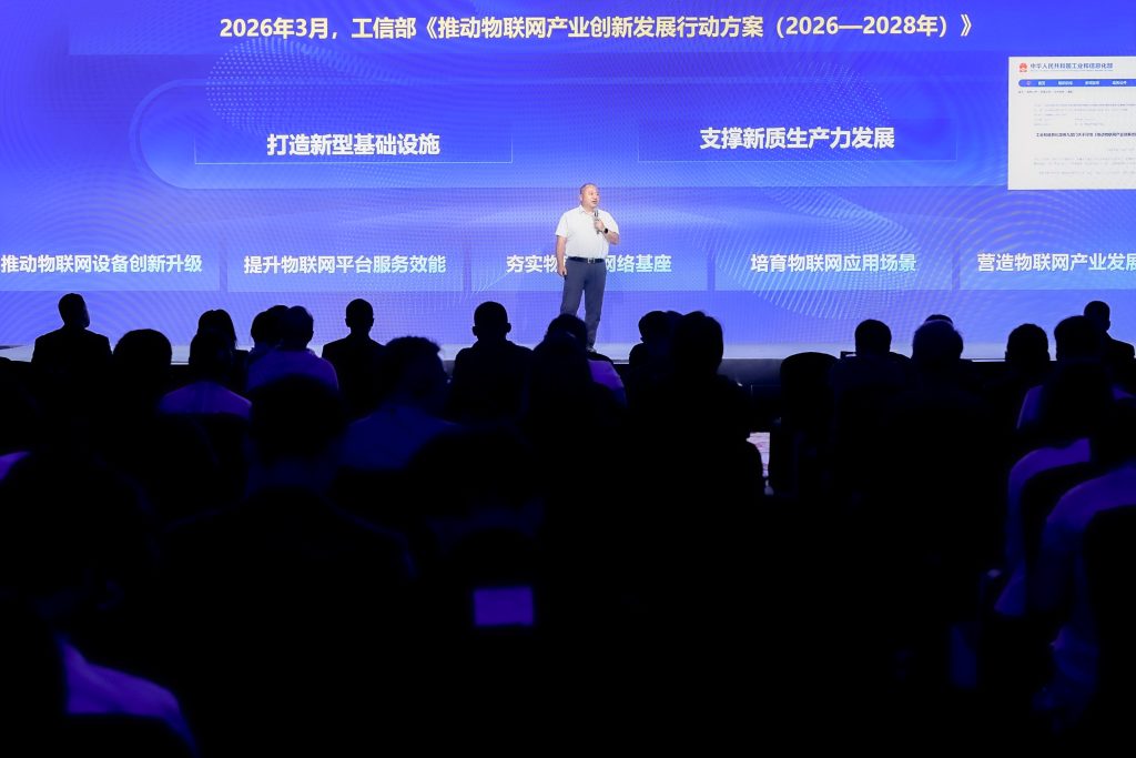 A man speaks on stage in front of a blue screen with Chinese text and white diagrams; an audience sits facing him in a darkened auditorium. The event appears focused on industrial development plans from 2026 to 2028.