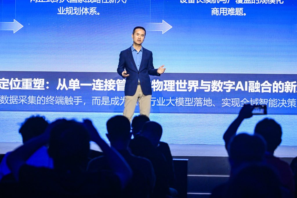A man in a blue blazer and khaki pants stands on a lit stage, speaking to an audience. The background features large Chinese and English text about AI, illuminated in blue. Silhouetted audience members are seen in the foreground, some raising hands.